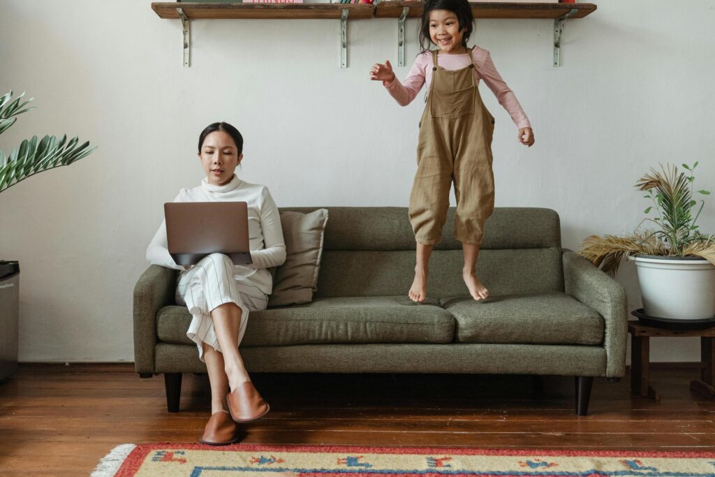 Mom on laptop with child jumping on couch next to her
